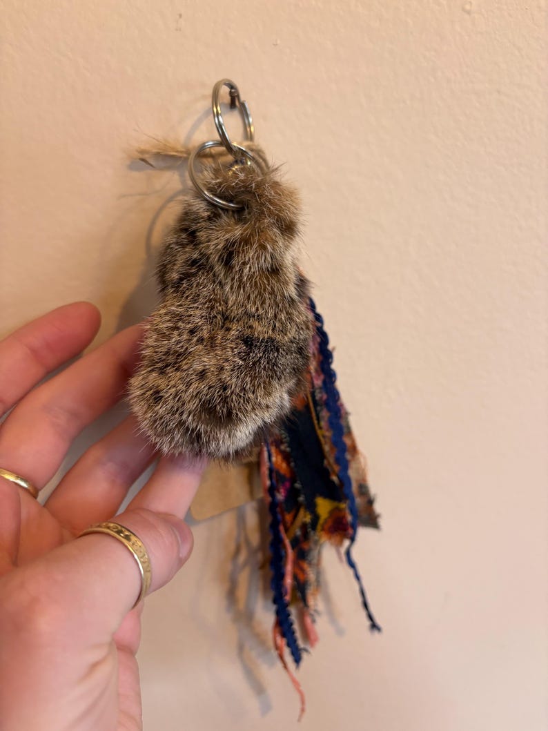 May include: A keyring featuring a fluffy, brown and black mottled fur charm attached to a silver metal ring. The charm is accented with a cluster of hanging fabric strips in navy blue, orange, and yellow floral patterns with frayed edges.