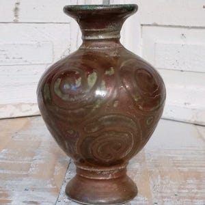 May include: A ceramic vase with a bulbous body and a flared rim, featuring a glossy brown glaze with swirling patterns in muted green and copper tones. The vessel has a distinct pedestal base and a smooth, reflective surface finish throughout.