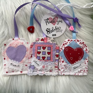 May include: Three fabric heart-shaped tags with pink, blue, and white patterns. The tags are decorated with lace and have the words "Heart", "Love", and "Heart" written on them. The tags are hanging from white ribbon and are attached to a white circle with the text "Thank you Shulakbee Shop" on it.
