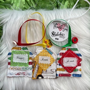 May include: Three fabric gift tags with a school bus and crayons design. The tags are labeled "Happy", "Teach", and "Read". The tags are attached to a white fluffy surface with a white string and a red button.