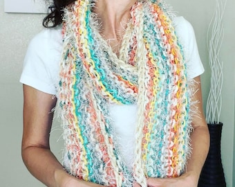 May include: A colorful, multi-colored, crocheted scarf with fringe. The scarf is made with a variety of yarns in shades of white, peach, teal, and yellow.