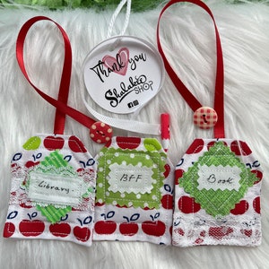 May include: Three red and white apple-themed fabric gift tags with white lace trim. The tags are labeled "Library", "BFF", and "Book".