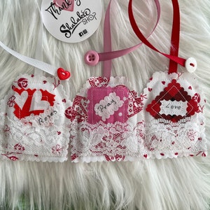 May include: Three handmade fabric heart-shaped ornaments with lace trim. The ornaments are decorated with red, pink, and white fabric and have the words "Roses", "Pray", and "Love" written on them. The ornaments are hanging from white and red ribbons.