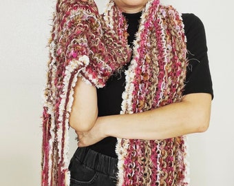 May include: A handmade, fringed scarf in shades of burgundy, pink, cream, and brown. The scarf has a textured, woven appearance with a fluffy, fringed edge. The scarf is wrapped around the neck and draped over the arm.