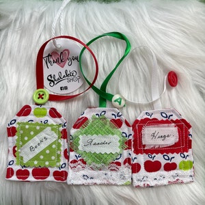 May include: Three red and white apple-themed gift tags with white lace trim. The tags are decorated with green polka dots and have handwritten messages: "Books", "Teacher", and "Hugs".