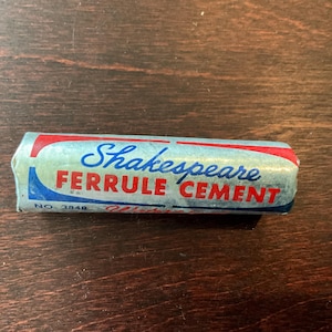 May include: A vintage Shakespeare Ferrule Cement tube, with a blue, red, and white design. The text "Shakespeare Ferrule Cement" is printed on the tube. The tube is cylindrical and appears to be made of paper or a similar material.