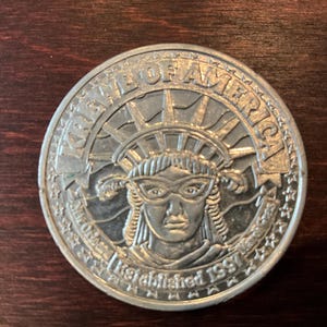May include: A silver-colored commemorative coin featuring the Statue of Liberty with the text "NEW ORLEANS" and "LAND OF AMERICA" around the edge. The coin is dated "Established 1997".