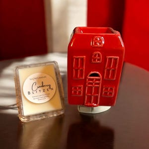 May include: A red ceramic house-shaped wax warmer with a white base. A square wax melt is in front of the warmer. The wax melt is labeled "Cranberry Blitzen" and is part of the "Winter Luxe Collection".