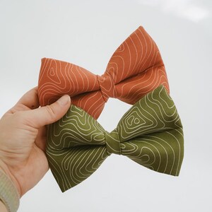 May include: Two bow ties, one burnt orange and one olive green, are held up against a white background. Both bow ties feature a white topographic map pattern. The bow ties are made of fabric.