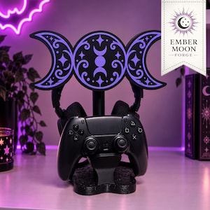 May include: A black gaming controller and headset stand featuring a triple moon symbol with purple accents and intricate scrollwork. The base is black with a textured pattern. A tag at the top displays the text Ember Moon Forge with a moon icon.