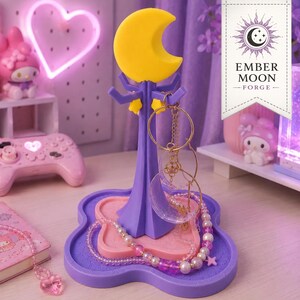 May include: A purple jewelry stand featuring a yellow crescent moon topper and a four-pointed base with a pink tray insert. The stand has multiple hooks for hanging accessories, with a beaded necklace and circular pendants draped around the central post.