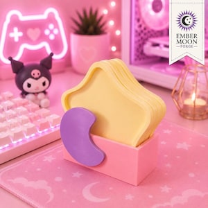 Puede incluir: Un juego de posavasos amarillos en forma de estrella guardados en un soporte rectangular rosa con un acento de luna creciente morada en el frente. Los posavasos están apilados verticalmente en el soporte sobre una superficie rosa con estrellas.