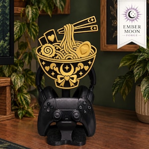 May include: A black and gold headphone and controller stand featuring a decorative ramen bowl topper with noodles, chopsticks, and heart motifs. The stand supports a black gaming controller and headphones, with the Ember Moon Forge logo visible.