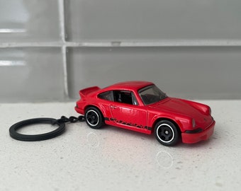 May include: A red toy car keychain with black wheels and a black key ring. The car has a spoiler and a black graphic on the side. The keychain is a miniature replica of a classic sports car.