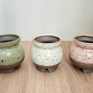 May include: Three ceramic pots with speckled glaze. The pots are in shades of green, white, and pink. Each pot has a brown base and three small feet.