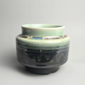 May include: A ceramic vessel with a wide, rounded body and a short, flared neck. The glaze features a gradient transition from a pale mint green at the top to a deep, glossy black at the base, with a band of blue and white drip-like accents.