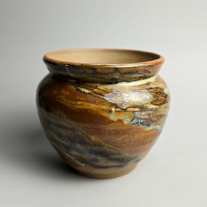 May include: A handmade ceramic pot with a wide, rounded body and a slightly flared rim. The pot features a blend of earthy tones, including browns, tans, and hints of blue, with a glossy finish. The pottery is likely used for decorative purposes.