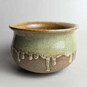 Peut inclure: Un petit pot en céramique brun avec un glaçage vert qui a coulé sur les côtés. Le pot a une forme arrondie et une large ouverture.
