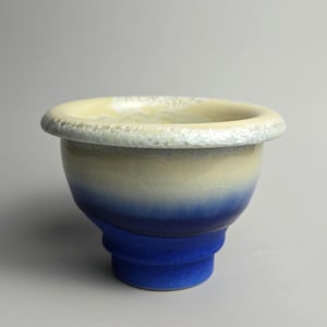 May include: A ceramic bowl featuring a gradient colour scheme that transitions from a deep blue base to a lighter cream-coloured upper section. The wide, rounded rim has a textured, crystalline white finish, and the entire surface displays a fine crackle glaze pattern throughout the exterior.