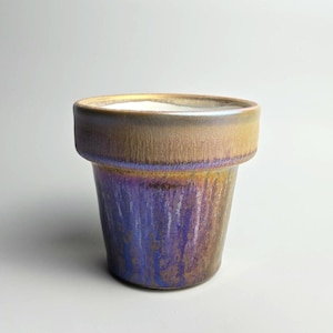 May include: A ceramic planter with a metallic finish. The rim is a warm brown, transitioning to a purple and gold body. The planter has a cylindrical shape with a slightly wider rim, suitable for small plants or succulents.