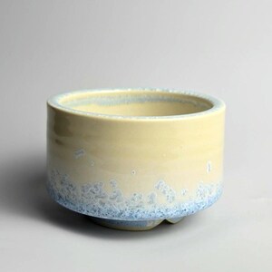 May include: A ceramic bowl with a pale yellow glaze and a blue crystalline pattern around the base. The bowl has a rounded shape and a smooth, glossy finish. The interior is a lighter shade of yellow.