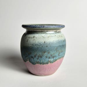 May include: A handmade ceramic vase with a rounded form and a gently widening rim. The vase displays a colour gradient, shifting from a pink base to teal, light blue, and a dark blue rim. Suitable for home decor.