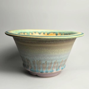 May include: A ceramic bowl featuring a wide, flared rim and a tapered base. The exterior displays a gradient of colors, transitioning from a soft pink at the bottom to light blue, beige, and a vibrant teal drip pattern along the upper edge.