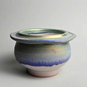 May include: A ceramic bowl with a wide, flat, iridescent rim that transitions from a metallic green to a soft gold. The rounded body features a gradient of blue and purple hues, fading into a solid white base with a smooth, glazed finish.