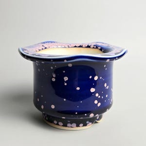May include: A deep blue ceramic planter with a wide, wavy rim and a speckled pattern of light pink dots. The interior of the pot is a smooth, light cream colour, and the base features a small, raised footed design with a visible light-coloured rim.