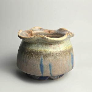 May include: A ceramic bowl with a ruffled, wavy rim and a rounded body. The glaze features a blend of muted earth tones, including beige, tan, and soft brown, accented by vertical streaks of deep blue that extend from the base towards the centre.