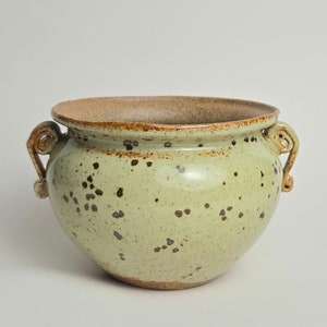 May include: A ceramic planter with a light green glaze and dark brown speckles. The rim and handles are a darker brown. The planter has two decorative, curled handles on either side.