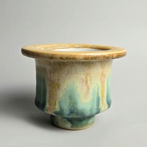 May include: A ceramic planter with a wide, flat rim and a tapered base. The exterior features a reactive glaze with blended tones of sage green, deep teal, and warm golden-brown, creating a fluid, mottled appearance across the curved surface.