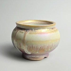 May include: A ceramic pot with a rounded body and a slightly flared rim. The pot has a cream-coloured base with iridescent gold, pink, and purple hues. The rim is a darker gold colour. The pot sits on a small, rounded base.
