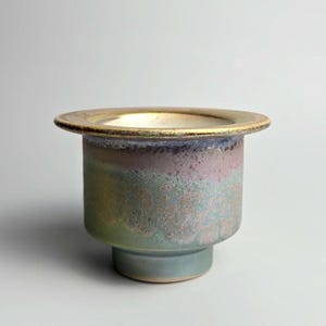 May include: A ceramic bowl with a wide, flat rim and a cylindrical body that tapers to a smaller base. The exterior features a mottled glaze with blended hues of sage green, dusty rose, and muted blue, while the interior rim has a metallic gold finish.
