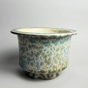 May include: A ceramic planter with a wide, flat rim and a footed base. The exterior features a crystalline glaze with a mottled pattern of soft blue, sage green, and white tones, while the interior is finished in a smooth, off-white glaze.