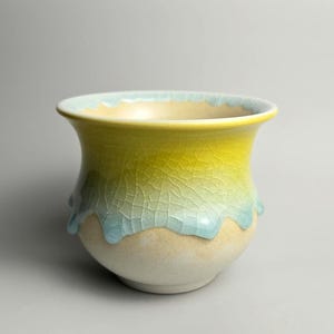 May include: A ceramic bowl with a flared rim, featuring a gradient glaze that transitions from a pale beige base to a vibrant yellow upper section, overlaid with a distinct blue crackle glaze pattern that drips down the sides of the vessel.