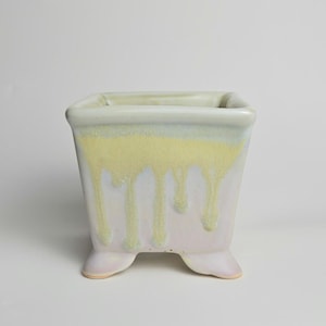 May include: A square ceramic planter with a light green and yellow glaze that drips down the sides. The planter has a pale pink base and sits on three small feet. The interior is a light green colour.