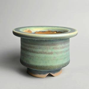 May include: A ceramic plant pot with a wide, rounded rim and a cylindrical body. The pot has a blend of green, brown, and blue tones, with a textured surface. The base has a unique, three-footed design. A decorative piece for plants.