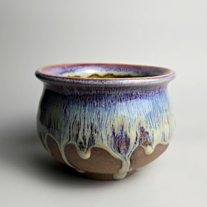 May include: A small ceramic bowl with a rounded shape and a wide, flared rim. The exterior features a reactive glaze with layers of deep purple, light blue, and cream colours that drip down over the exposed, unglazed brown clay base.