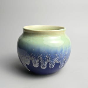 May include: A ceramic bowl with a rounded shape and a wide opening. The exterior features a gradient colour scheme, transitioning from a pale cream at the rim to a light green, then to a deep blue at the base, with a white speckled drip pattern.