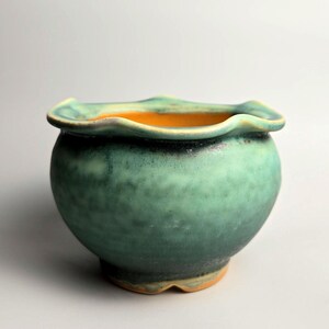 May include: A small ceramic bowl with a rounded body and a wavy, scalloped rim. The exterior features a mottled, matte finish in shades of teal and dark green, while the interior and the base reveal a contrasting warm, bright orange glaze.