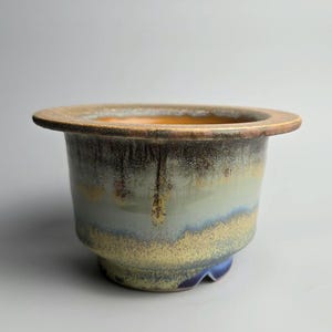 May include: A ceramic planter with a wide, flat rim and a tapered body. The exterior features a reactive glaze with blended tones of dark brown, sage green, and pale yellow, while the interior is finished in a smooth, solid warm orange glaze.