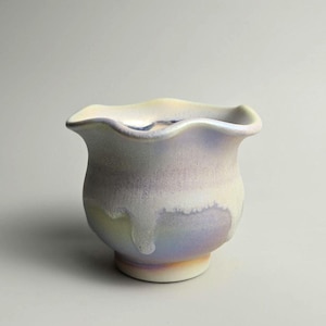 May include: A ceramic vase with a frilled rim and a rounded body. The vase has a pearlescent finish with shades of lavender, cream, and pale yellow. The base is a soft beige colour, and the design is elegant and artistic.
