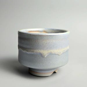 May include: A ceramic tea bowl with a cylindrical shape and a footed base. The exterior features a matte, light blue-grey glaze with a textured, cream-coloured drip pattern around the upper section. The interior rim reveals a warm, orange-toned clay.