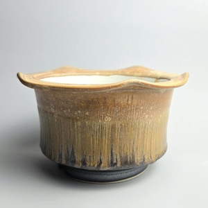 May include: A ceramic bowl with a wide, wavy rim and a tapered base. The exterior features a gradient glaze transitioning from a light tan at the top to a dark, charcoal-black at the bottom, with vertical drip patterns throughout the surface.