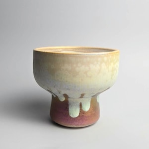 May include: A ceramic footed bowl featuring a wide, rounded upper section and a narrower base. The glaze transitions from a pale, creamy yellow at the top to a light blue drip pattern, ending in a deep, metallic reddish-brown finish at the bottom.