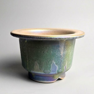 May include: A ceramic bowl with a wide, flat rim and a tapered base. The exterior features a multi-coloured iridescent glaze with dripping patterns in shades of metallic green, blue, and purple, transitioning to a soft golden hue at the top rim.