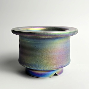 May include: A ceramic vessel with a wide, flat rim and a rounded body. The exterior has a textured, iridescent finish with hues of blue, green, and purple. The base has a small, decorative foot.