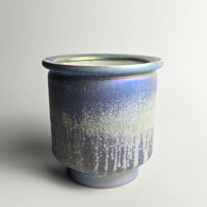 May include: A ceramic vase with a cylindrical form and a flared rim. The vase has a blue and iridescent finish with a textured, dripped effect on the lower half. The base is a lighter shade of blue.