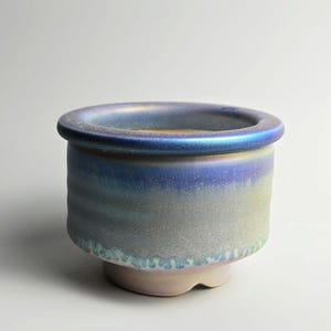 Peut inclure: Petit pot de fleurs rond en céramique avec une finition irisée texturée. Le pot présente un bord bleu et un dégradé de teintes bleues, vertes et violettes. La base est de couleur claire et neutre.